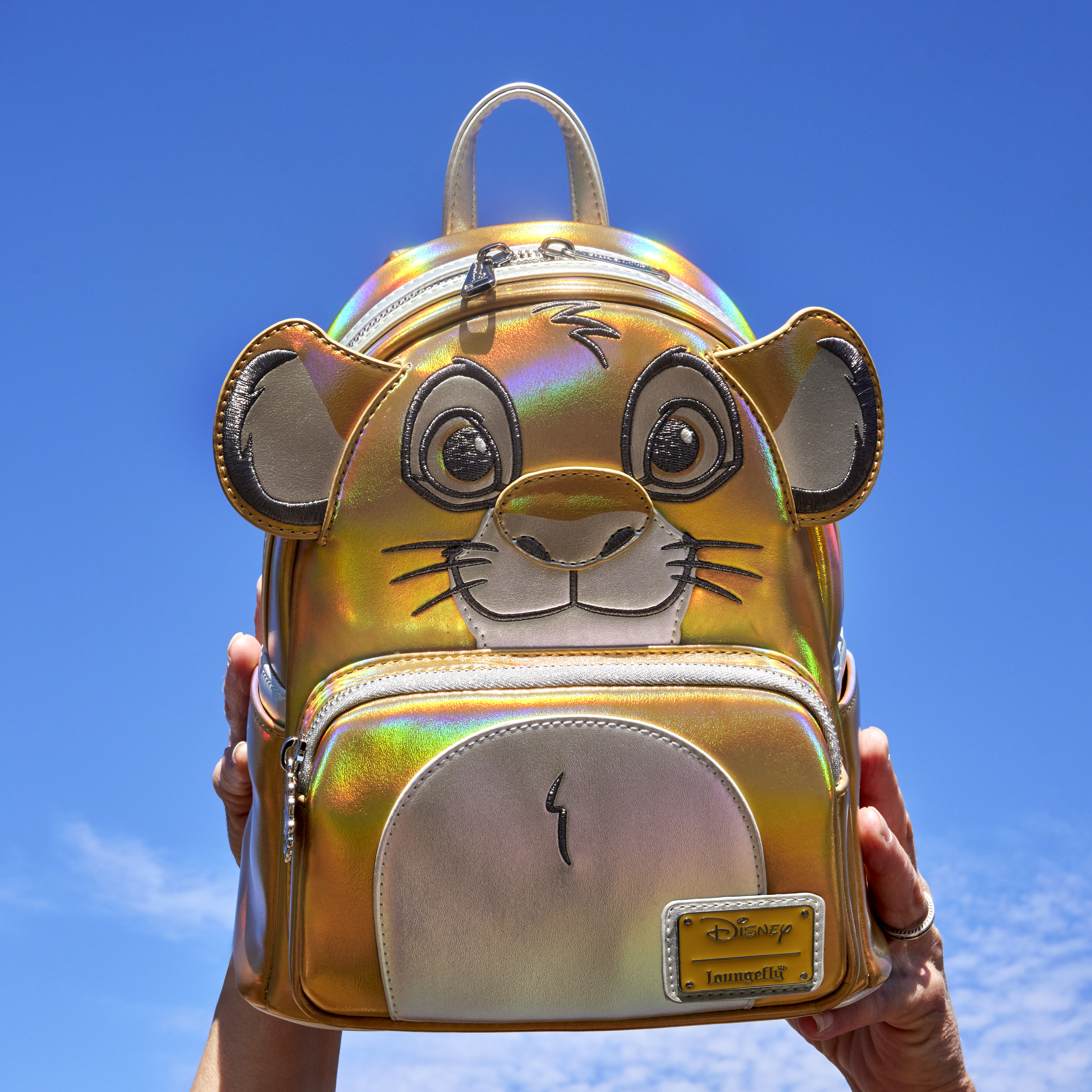 A pair of hands holds the Loungefly Disney100 Platinum Simba Cosplay Mini Backpack against a blue cloudy sky. The backpack shows Simba's lion cub face with shiny metallic details.