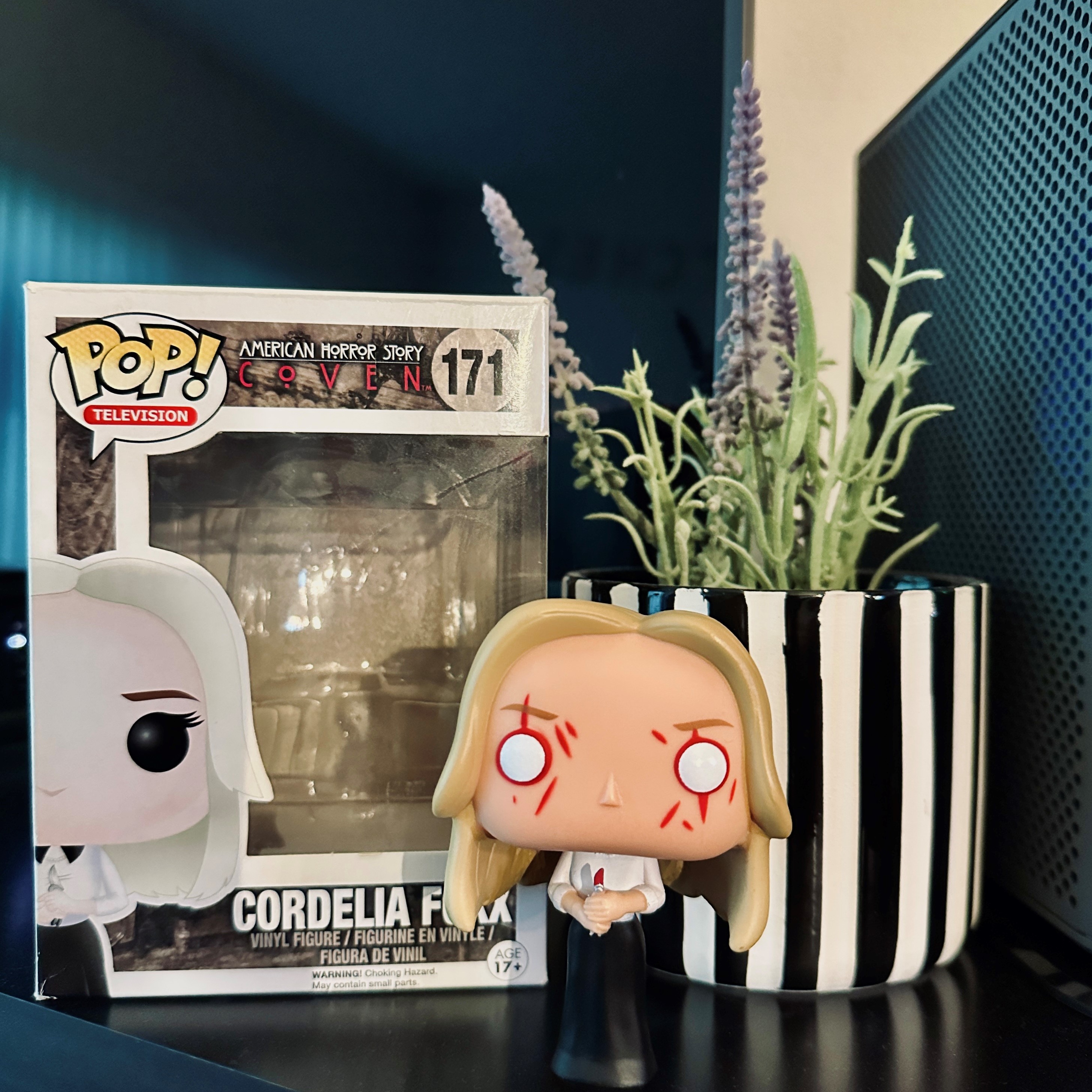 Pop! Cordelia Foxx, set on a stand, in front of her Pop! box and a plant in a black-and-white-striped vase.