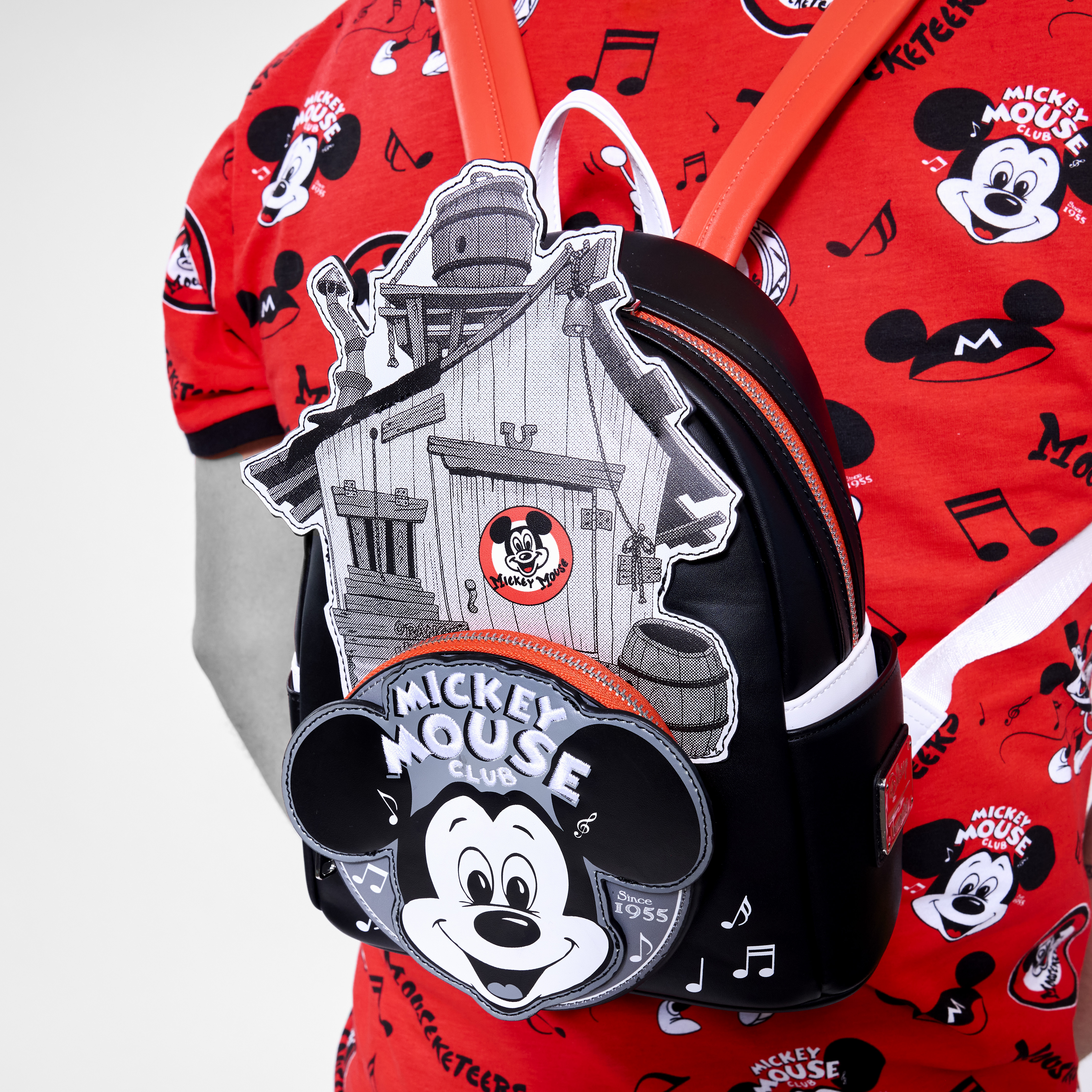 A man wears the Loungefly Disney100 Mickey Mouse Club Mini Backpack on his back. The backpack shows the Micky Mouse Clubhouse from the original TV series. The backpack appears against a red tee-shirt featuring an allover print of Mickey Mouse motifs.