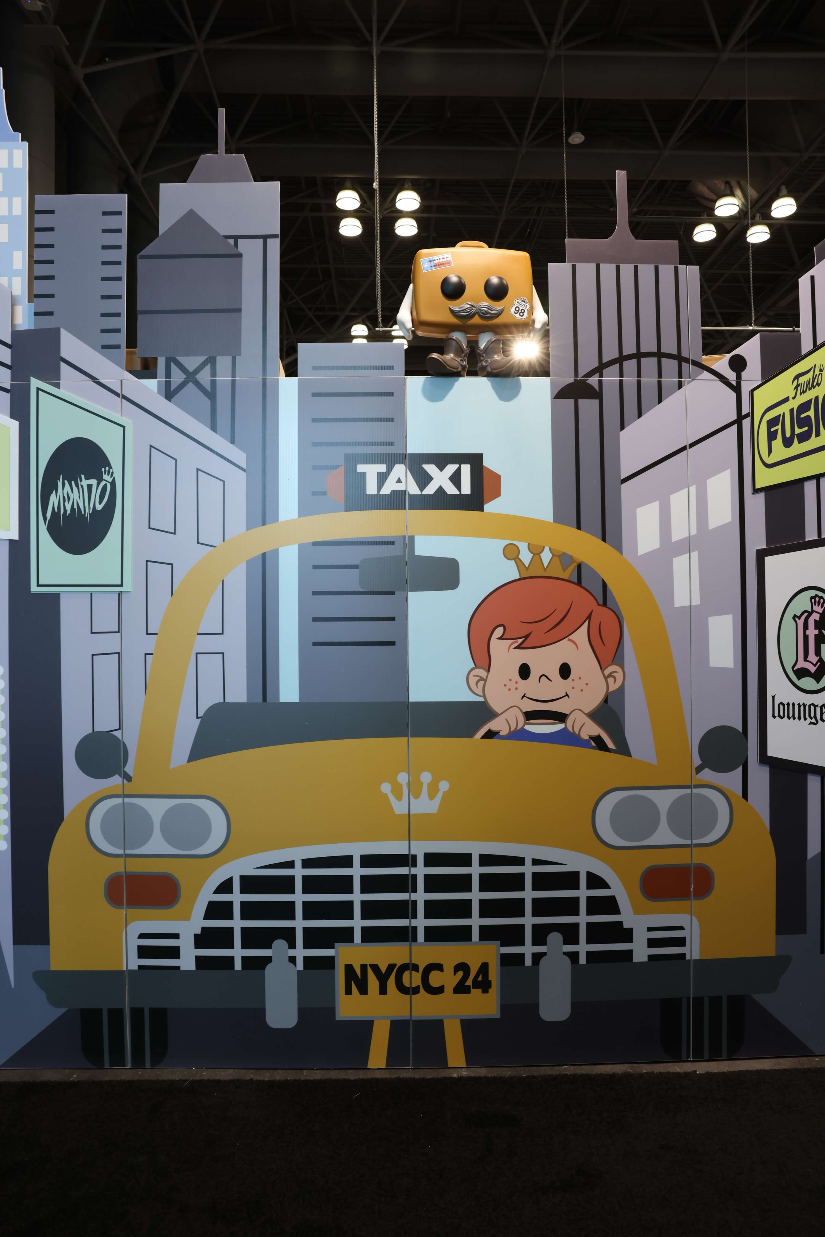"City" view of the booth with a cutout of Freddy Funko driving a taxi with high-rise buildings in the background.